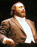 A man in a dress coat and white shirt opens his mouth, which is framed by dark facial hair.