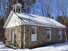 Ein kleines steinernes Gebäude im Winter. Schnee bedeckt den Boden und das Dach mit dem Backsteinkamin und einem kleinen offenen Glockenturm