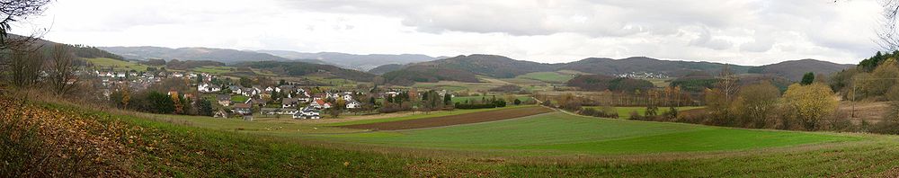 Blick auf Holzhausen am Hünstein (linke Bildhälfte) und das Dautphetal. Im Hintergrund (v.l.n.r.) der Nimerich (Breidenbacher Grund, 533 m), die Sackpfeife (674 m) und ihre Vorhöhen (u. a. Arennest, 592 m) und davor der Ort Dautphe; in der rechten Bildhälfte die westlichen Damshäuser Kuppen mit Hornberg (451 m), Eichelhardt (465 m, teils verdeckt), Schweinskopf (473 m, hinter Herzhausen) und Dusenberg (457 m)