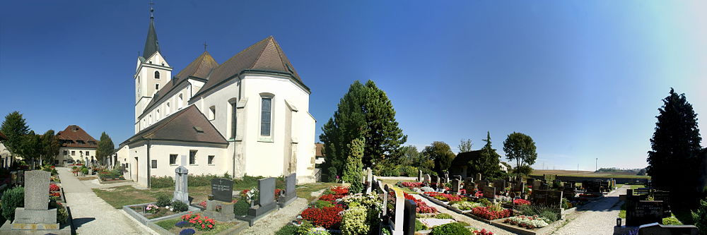 Panoramaansicht der Pfarrkirche Kleinhain umgeben vom örtlichen Friedhof: links das Klöster St.Josef, danach die Pfarrkirche, rechts davon die Aufbahrungshalle, ganz rechts das Friedhofskreuz. Rechts im Hintergrund einige Windräder des Windparks Kleinhain.