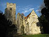 Auf der linken Seite befindet sich ein Kirchturm, in der Mitte die Ruine einer Mauer und rechts das Ende einer steinernen Kapelle mit einem roten Ziegeldach.