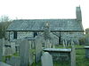 Die nördliche Seite einer steinernen Kirche mit Schieferdach in einem Friedhof; es gibt zwei Fenster, eine durchgehende Sakristei und ein Glockenstuhl auf der rechten Seite.