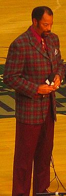 A man, wearing red and green jacket with a red shirt and tie, is standing on a basketball court while holding a microphone.