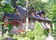 Ein hellbraunes Haus mit einem dunklen Dach in einer bewaldeten Umgebung.