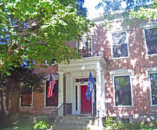 Ein Backsteingebäude mit einer roten Tür hinter einer Veranda. Die Flaggen der Vereinigten Staaten und New Yorks wehen hinter einem Baum auf der linken Seite