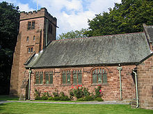 Eine Kirche mit einem mit Zinnen versehenen Turm auf der linken Seite, Rosen und eine Rasenfläche sind im Vordergrund zu sehen, Bäume stehen links und im Hintergrund