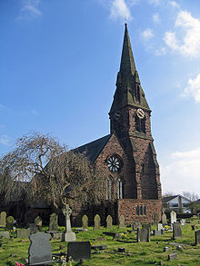 Kirchenfassade mit einer Rosette und dem steilen Dach einer Kirch im gotischen Stil. Im Vordergrund sind Grabsteine zu stehen, ein blattloser Baum steht auf der linken Seite.