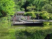 In einem kleinen See liegt einen Holzplattform, umgeben von Laternen, im Hintergrund eine Hecke und ein Wartehäuschen