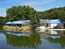 Ein Café erbaut aus gläsernen Gewächshäusern, am Ufer mit gelben Tischdecken und blauen Sonnenschirmen an den Tischen im Freien
