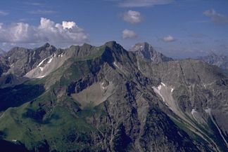 Zwölferkopf (der unscheinbare Berg im rechten Bildbereich im vorderen Bergzug) von Nordosten. In Bildmitte der grasige Gipfel des Elferkopfes. Rechts davon der Felsgipfel des Großen Widdersteins (scheinbar gleich hoch). Links davon der Felsgipfel des Liechelkopfes (berührt scheinbar die Wolken)