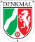 Schildförmige Denkmalplakette des Landes Nordrhein-Westfalen mit Wappen des Landes Nordrhein-Westfalen, darüber in Großbuchstaben der Schriftzug „Denkmal“, oben links und rechts sowie unten mittig ein Nagel