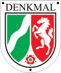 Schildförmige Denkmalplakette des Landes Nordrhein-Westfalen mit Wappen des Landes Nordrhein-Westfalen, darüber in Großbuchstaben der Schriftzug „Denkmal“, oben links und rechts sowie unten mittig ein Nagel.